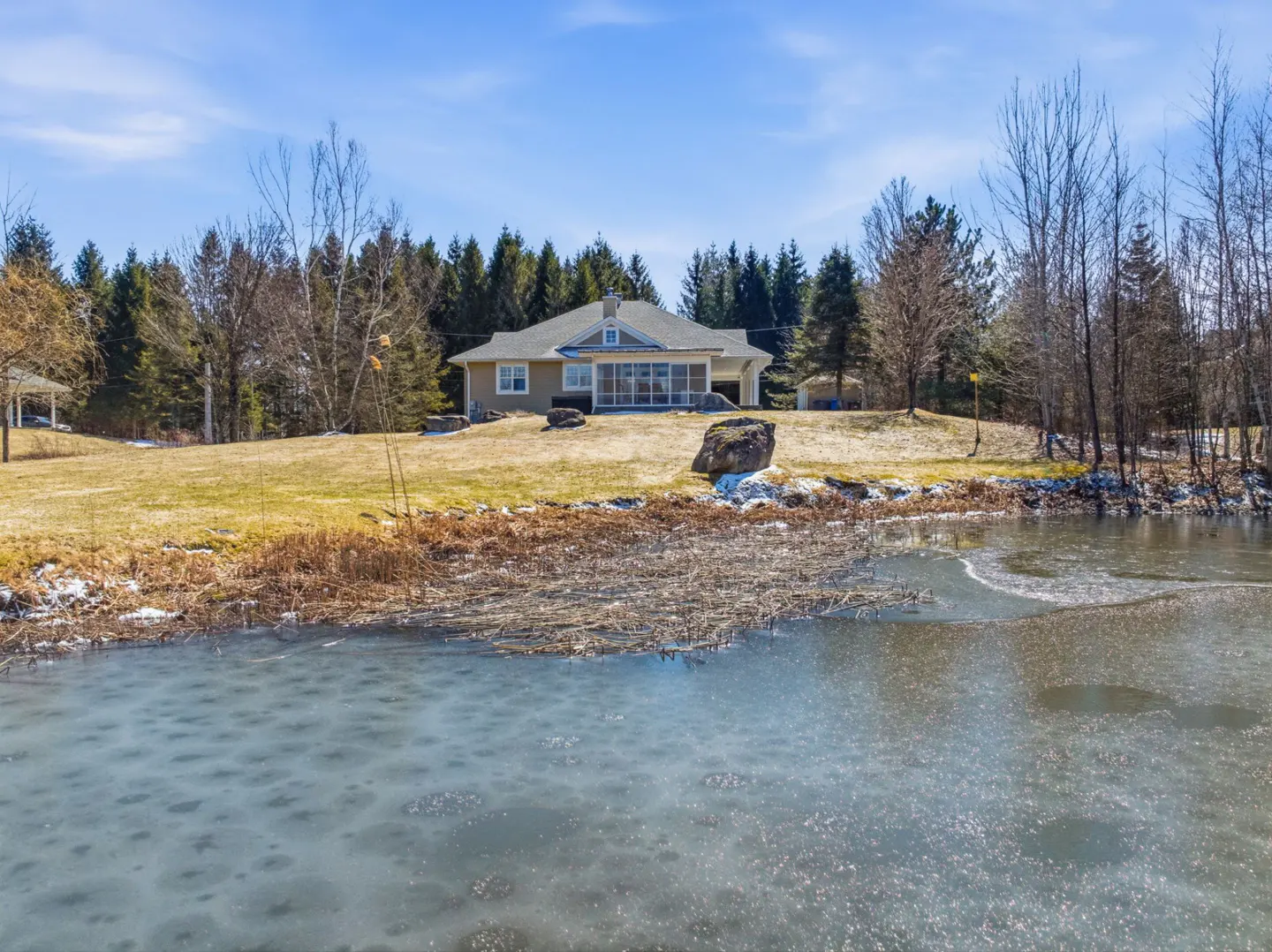 Tan house with screened porch overlooks a partially frozen pond. Trees line the property. Blue sky.