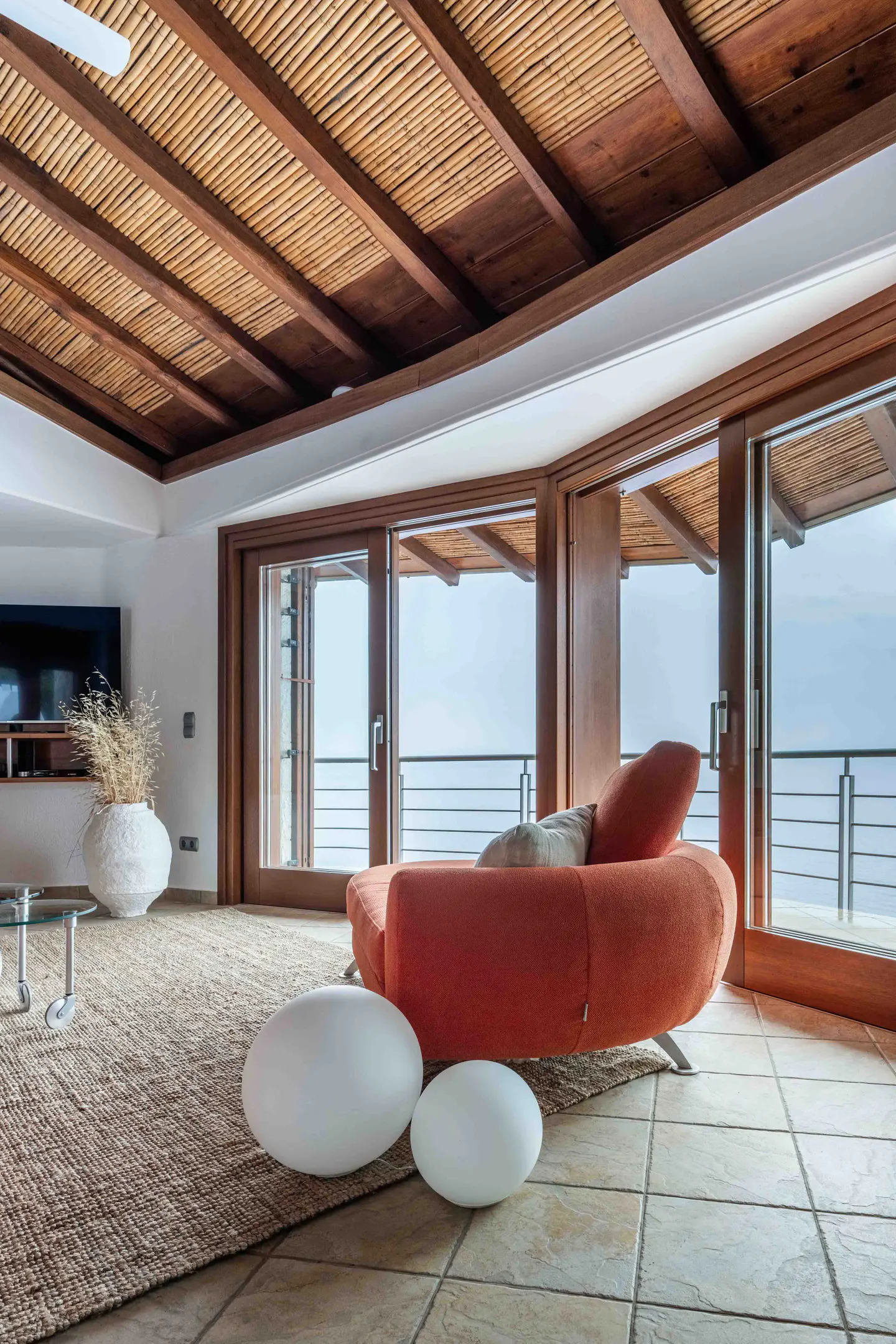 Living room with a bamboo ceiling, orange sofa, and large windows overlooking a balcony with an ocean view.
