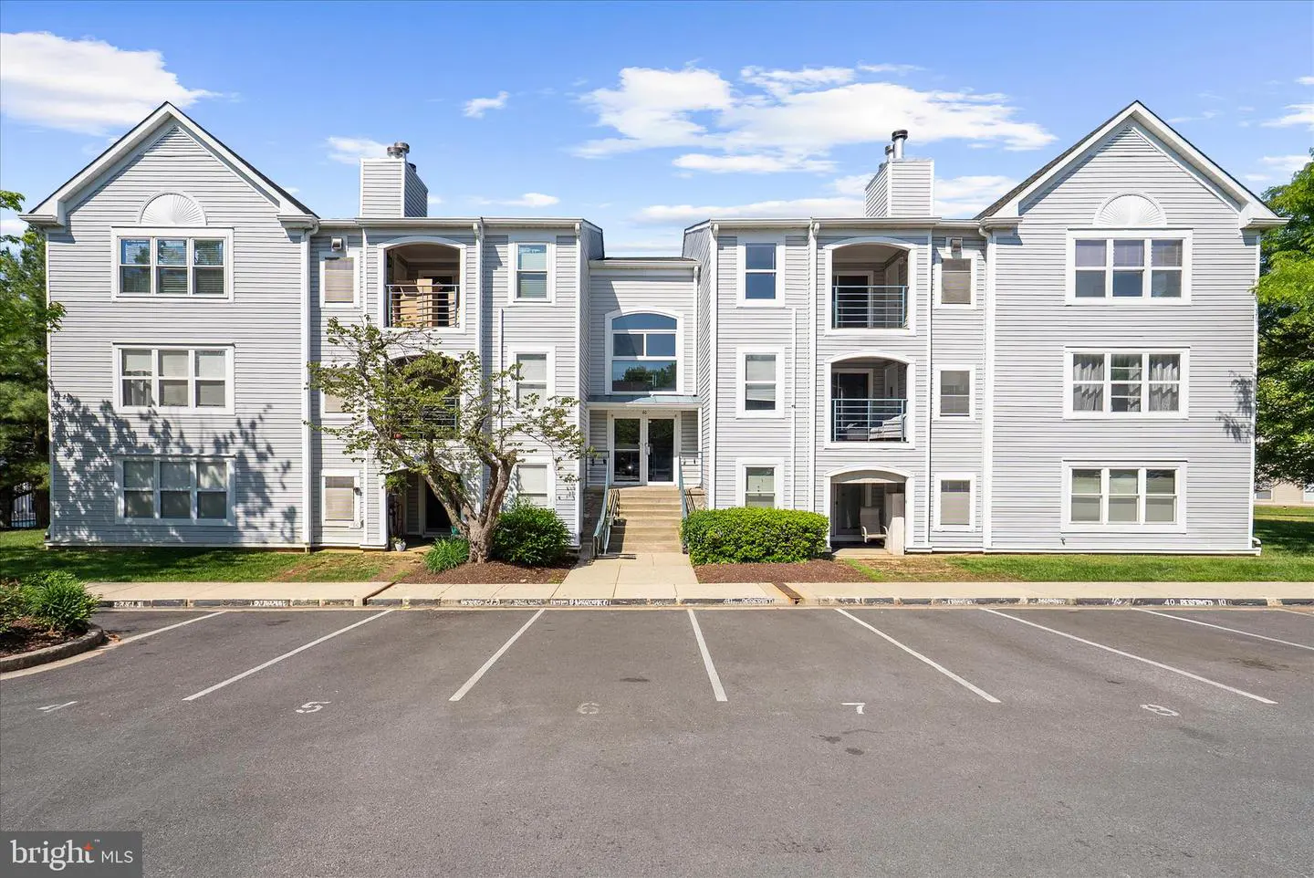 Exterior view of a light gray, three-story apartment building with balconies and a parking lot in front.