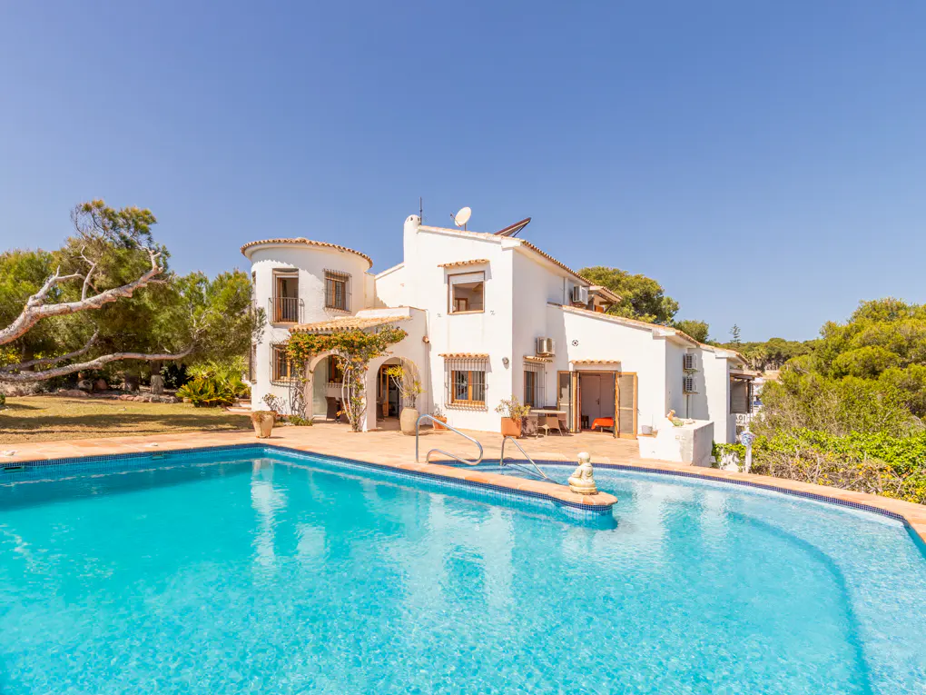 A white villa with a turquoise pool in the foreground under a clear blue sky.
