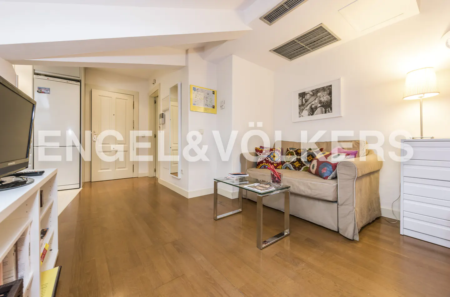 Living room with wood floors, beige sofa with colorful pillows, glass table, and white walls. Engel & Völkers logo overlay.