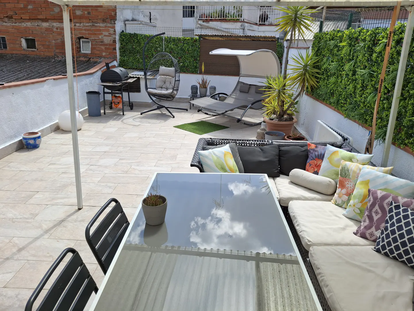Rooftop terrace with a glass dining table, cushioned sofa, hanging egg chair, lounge bed, grill, and green privacy walls.