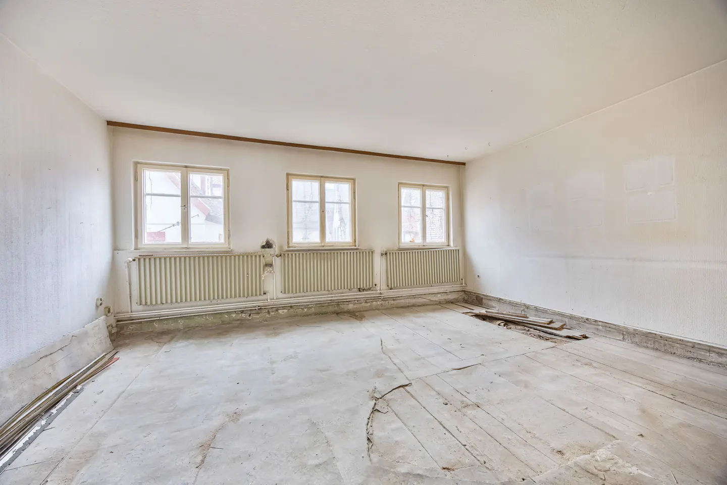 Empty room with white walls, three windows, and radiators. The floor is damaged and under construction.