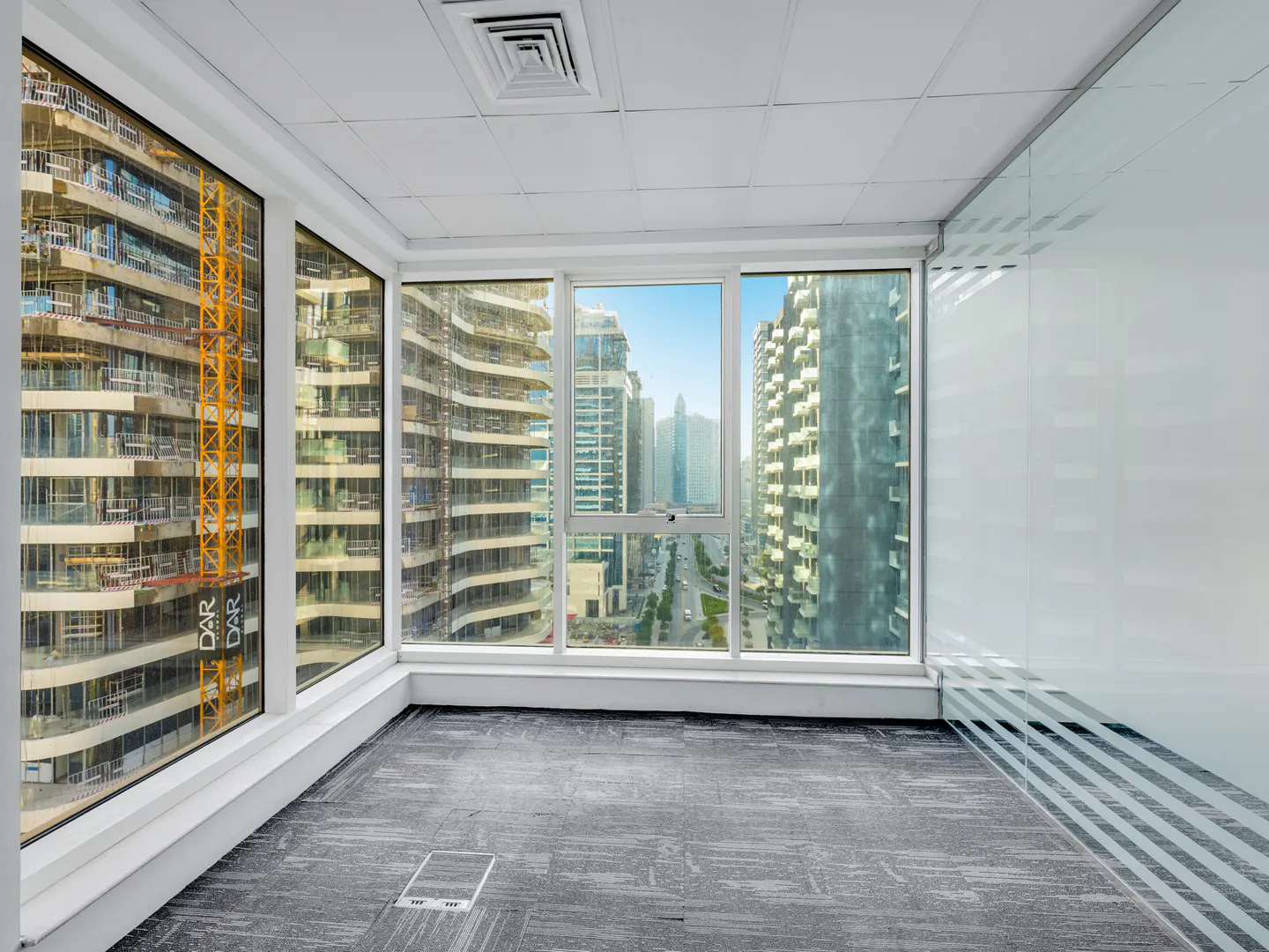 Empty office space with gray carpet and large windows. Outside, city views include buildings and a construction crane.