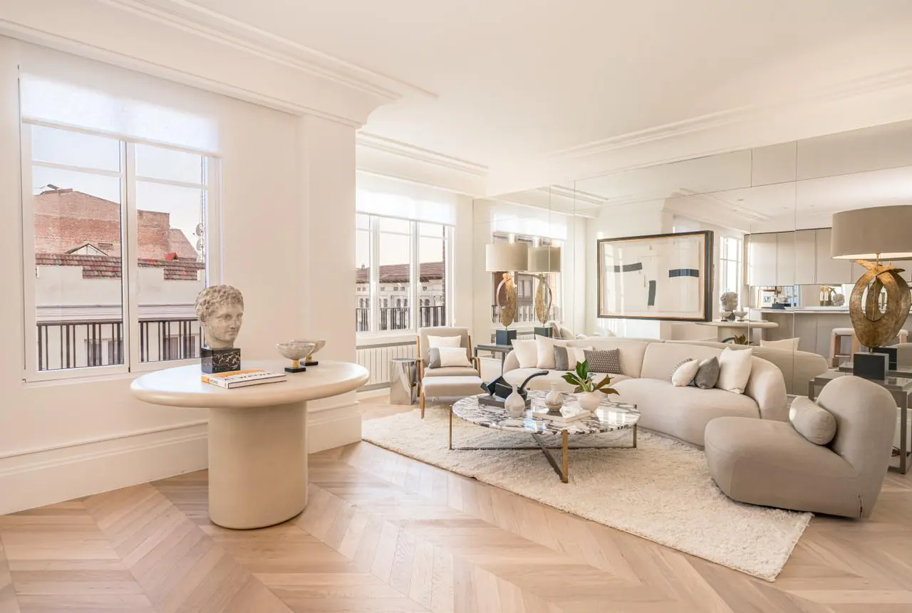 Bright, modern living room with white walls, herringbone floors, and large windows. A cream sofa, marble table, and bust statue add elegance.