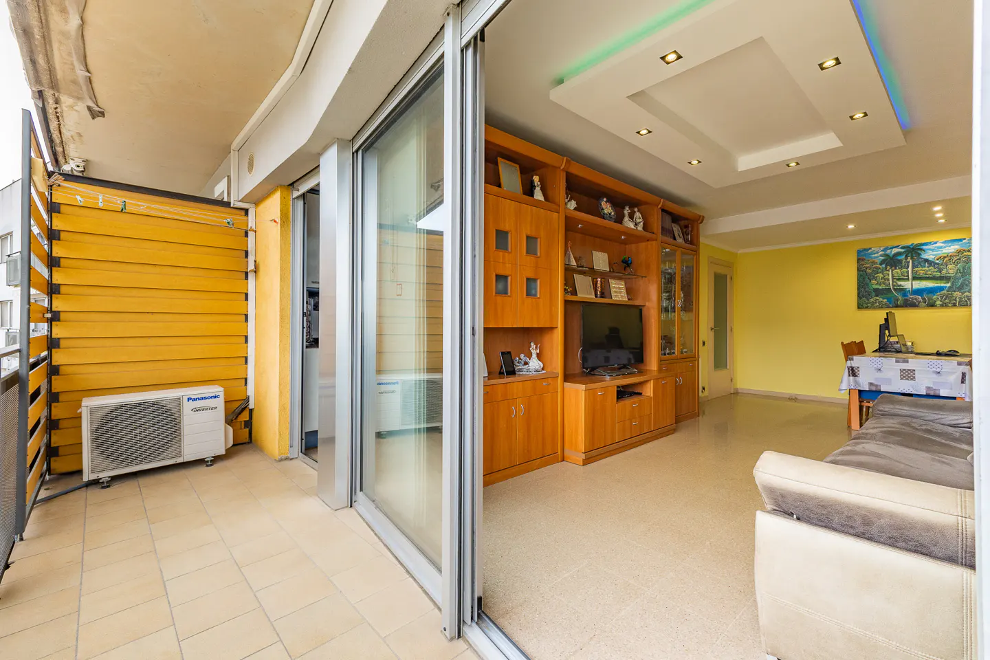 A bright living room with a balcony. The balcony has yellow slats and an AC unit. The living room has a wooden entertainment center and yellow walls.