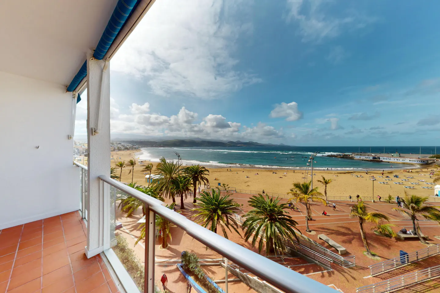 View from a balcony with a glass railing overlooking a sandy beach with palm trees and the ocean under a blue sky.
