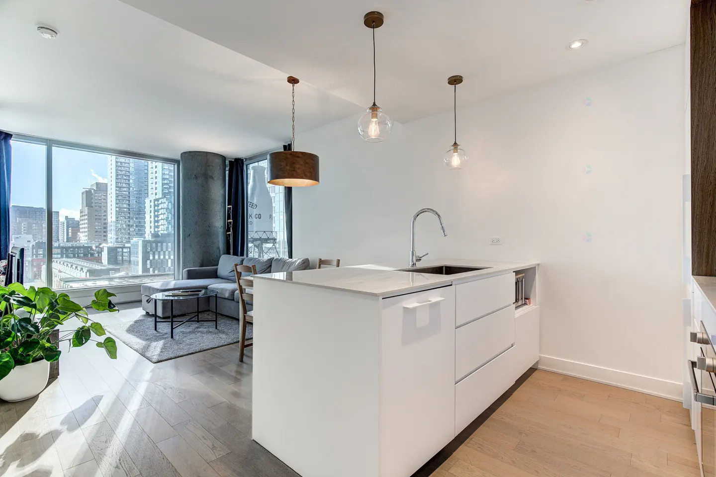 Modern kitchen with white island and sink, overlooking a living area with a grey sofa and floor-to-ceiling city windows.