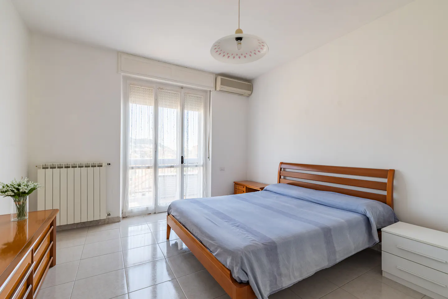 Bright bedroom with a wood-framed bed, blue striped bedding, white walls, and tiled floor. A dresser and nightstand are visible.