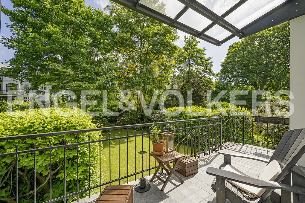 Balcony view with black metal railing, wooden chairs, and table. Lush green trees and lawn in the background. Overhead glass canopy.
