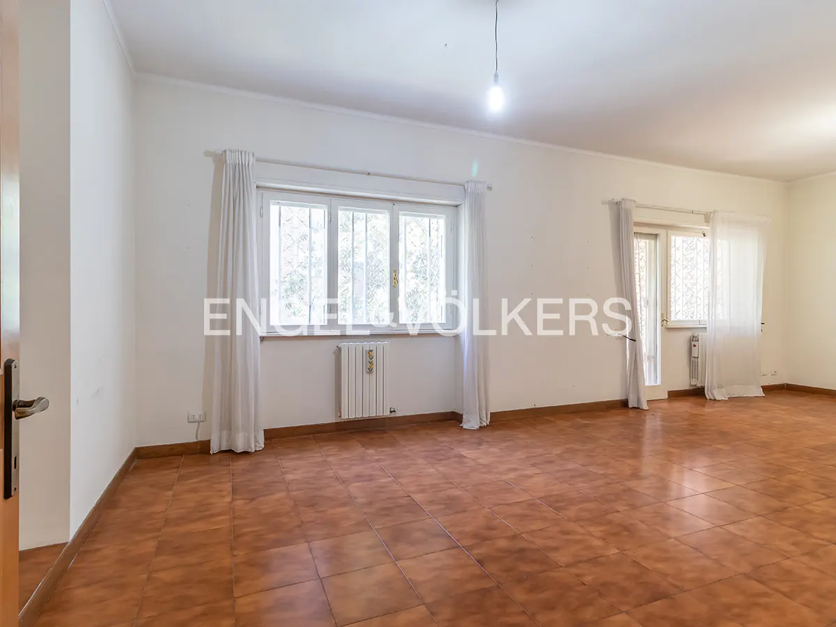 Empty room with white walls, brown tiled floors, large windows with white curtains, and a radiator.