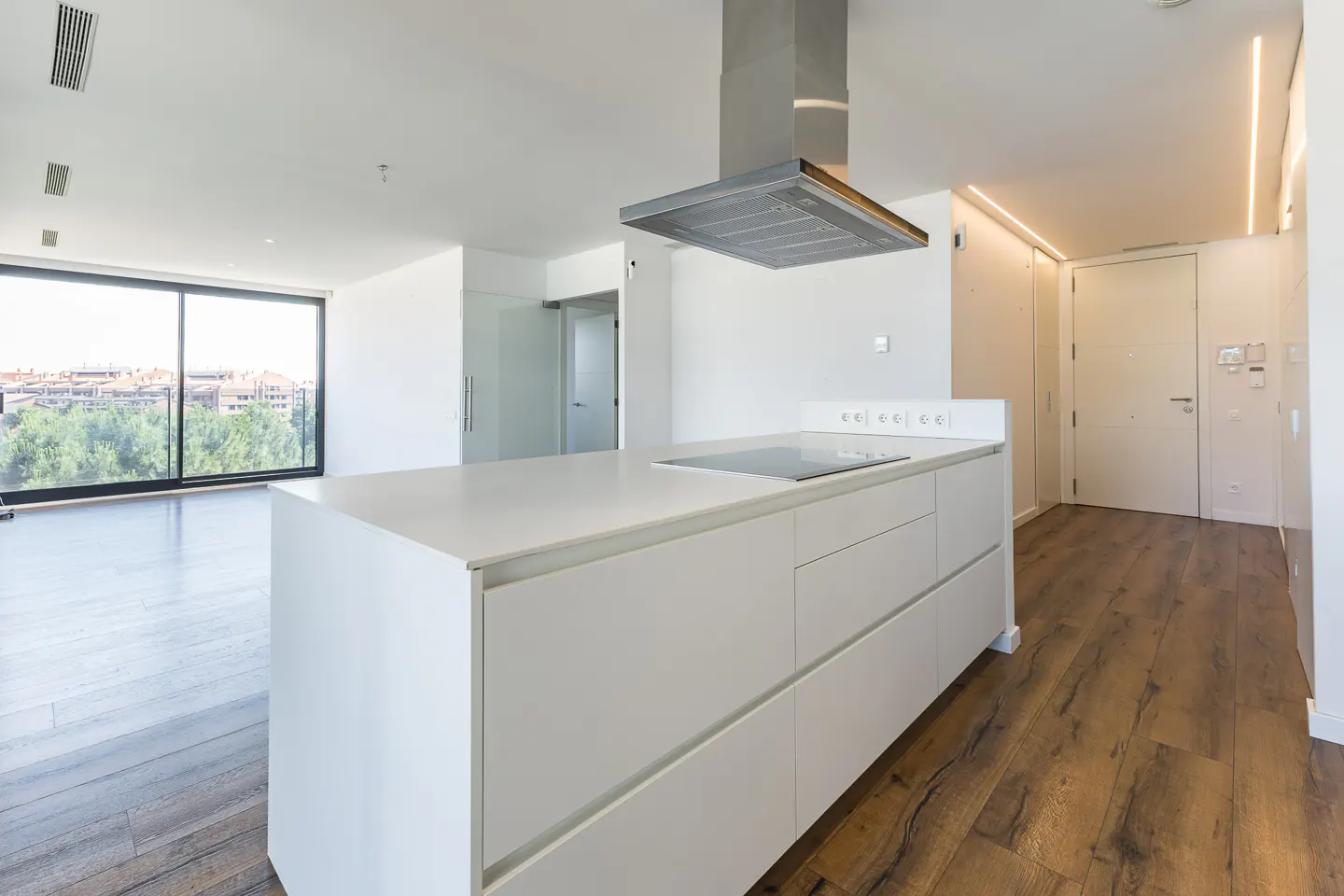 Bright, modern kitchen with white cabinets, island, and stainless steel range hood. Wood floors and large window with city view.