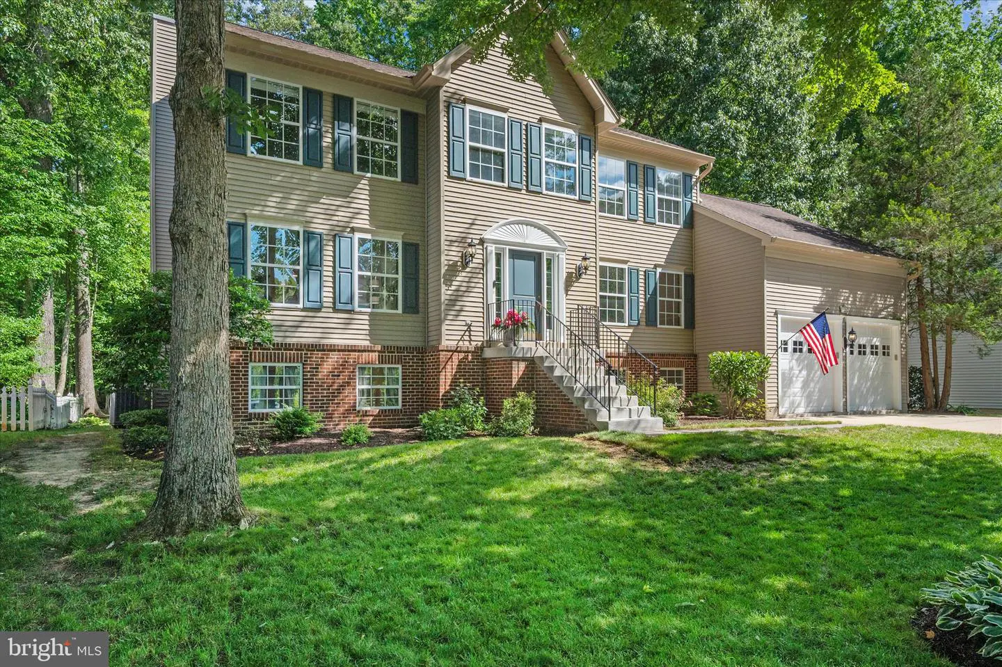 Two-story tan colonial home with dark shutters, brick foundation, front steps, and a two-car garage with an American flag.