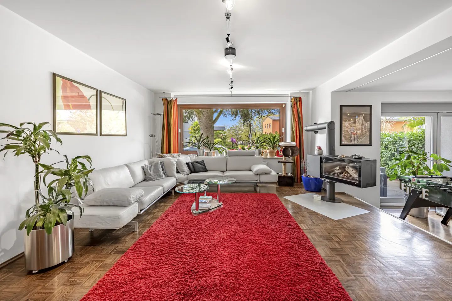 Bright living room with a red rug, white sofa, fireplace, and foosball table. Large window with orange curtains.