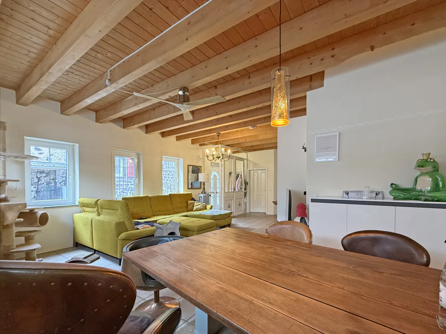 Living room with wood-beamed ceiling, yellow sectional sofa, and a large wooden table with brown leather chairs.