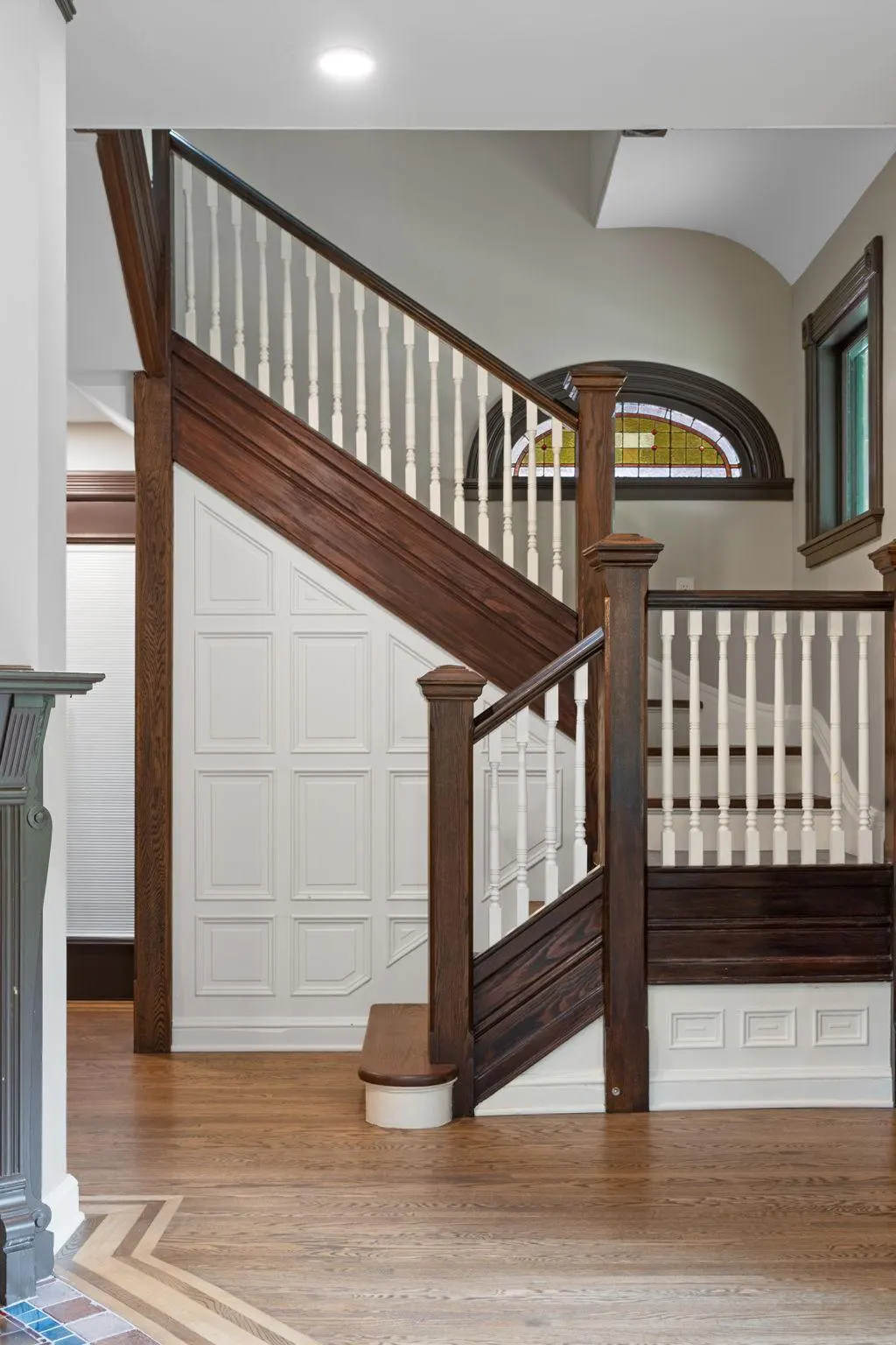 Interior view of a staircase with dark wood trim, white balusters, and paneled walls. Hardwood floors and a decorative window are visible.