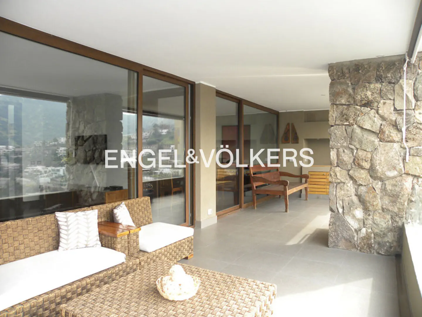 Modern covered terrace with wicker furniture, stone accent wall, and large glass sliding doors overlooking a mountain view.