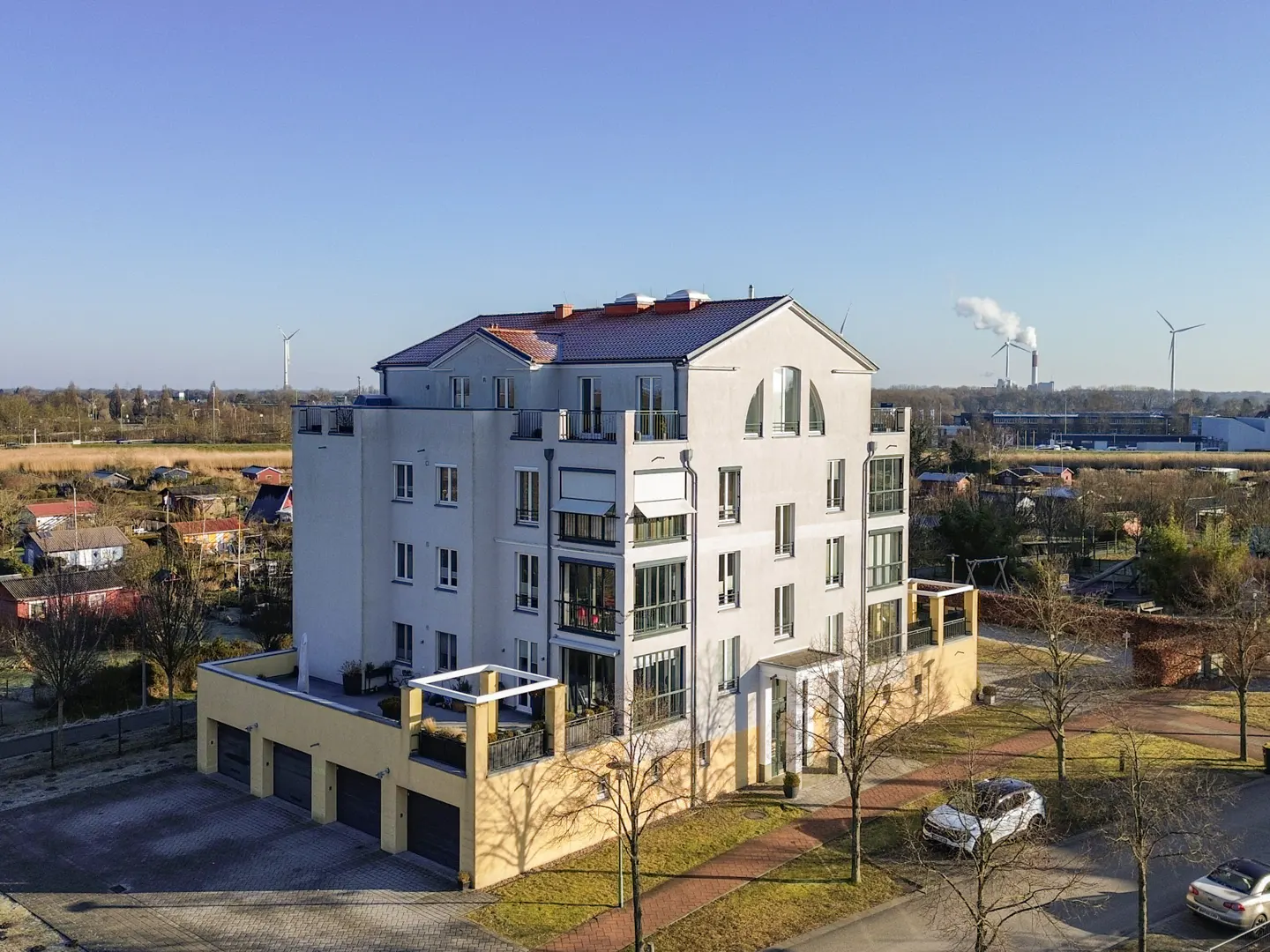 Modern apartment building with balconies, a yellow garage, and cars parked nearby on a sunny day. Wind turbines in the background.