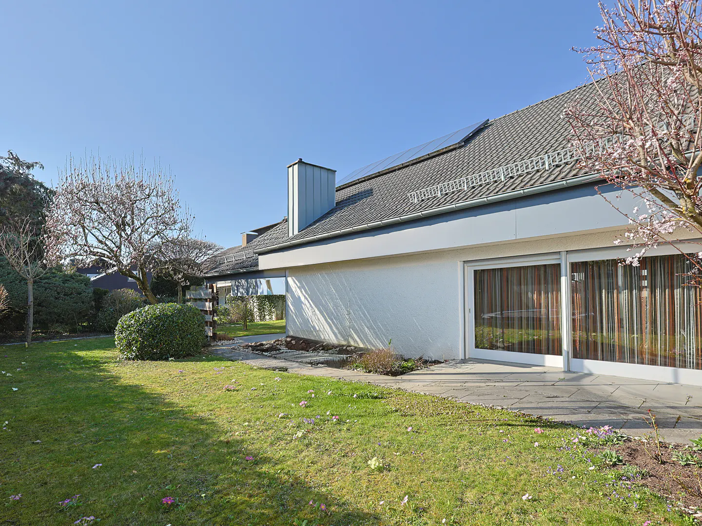 Exterior view of a one-story white house with a gray roof, solar panels, and a green lawn with trees and flowers.
