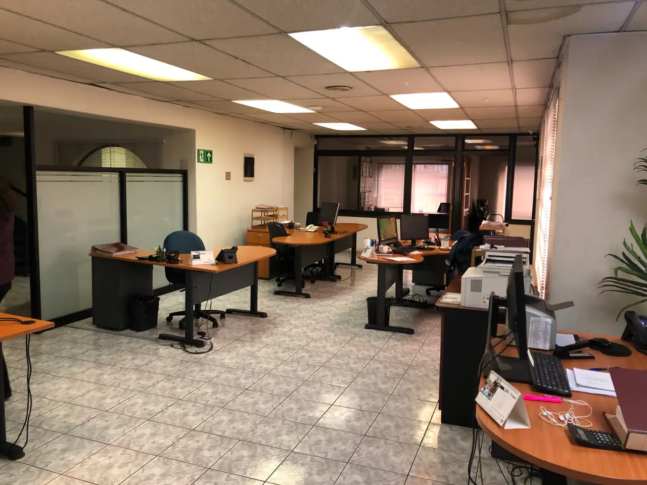 A bright, open-plan office with multiple wooden desks, black chairs, tiled floors, and fluorescent ceiling lights.