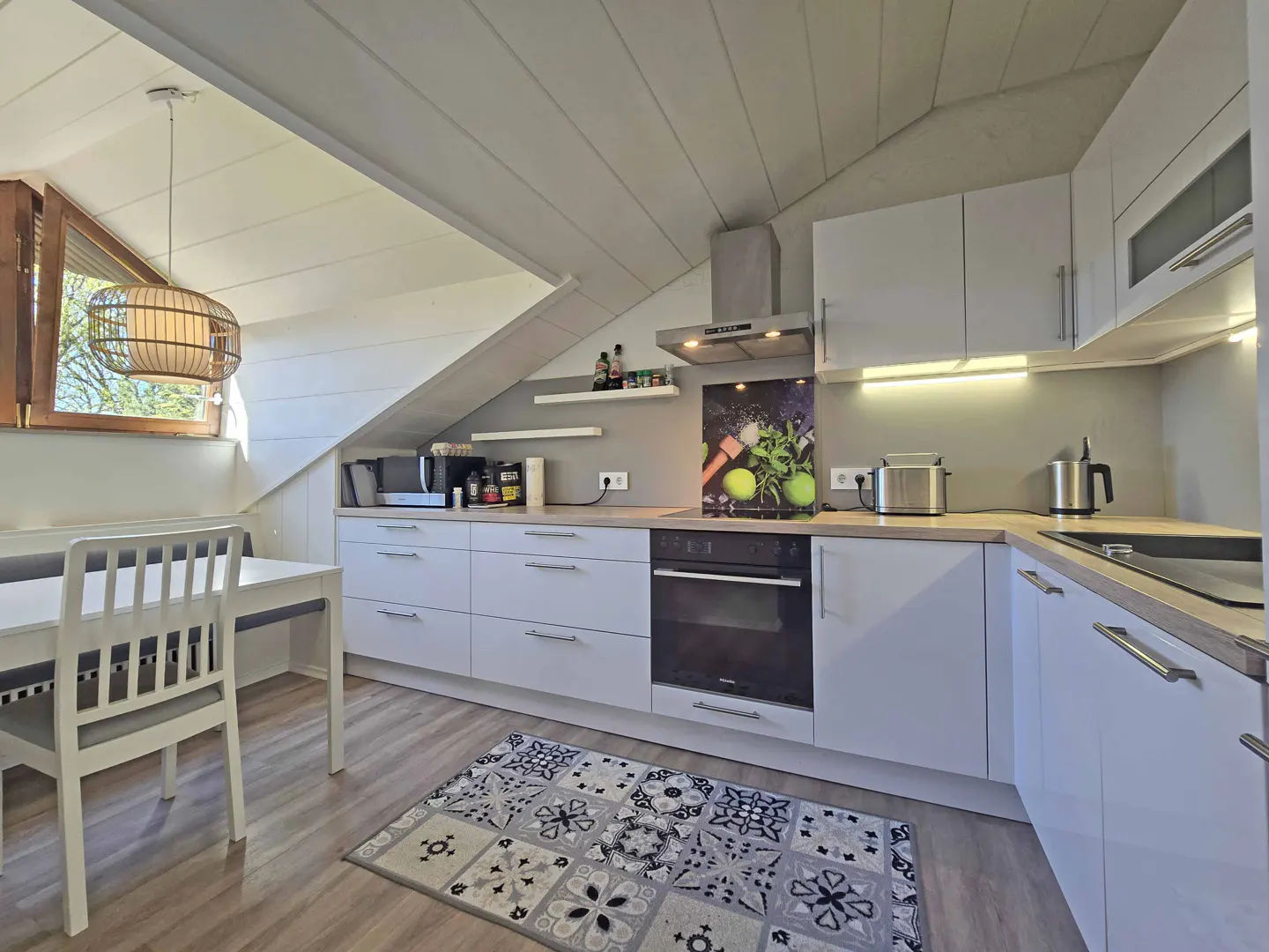 Modern attic kitchen with white cabinets, light wood countertops, a patterned rug, and a dining area under a sloped ceiling.