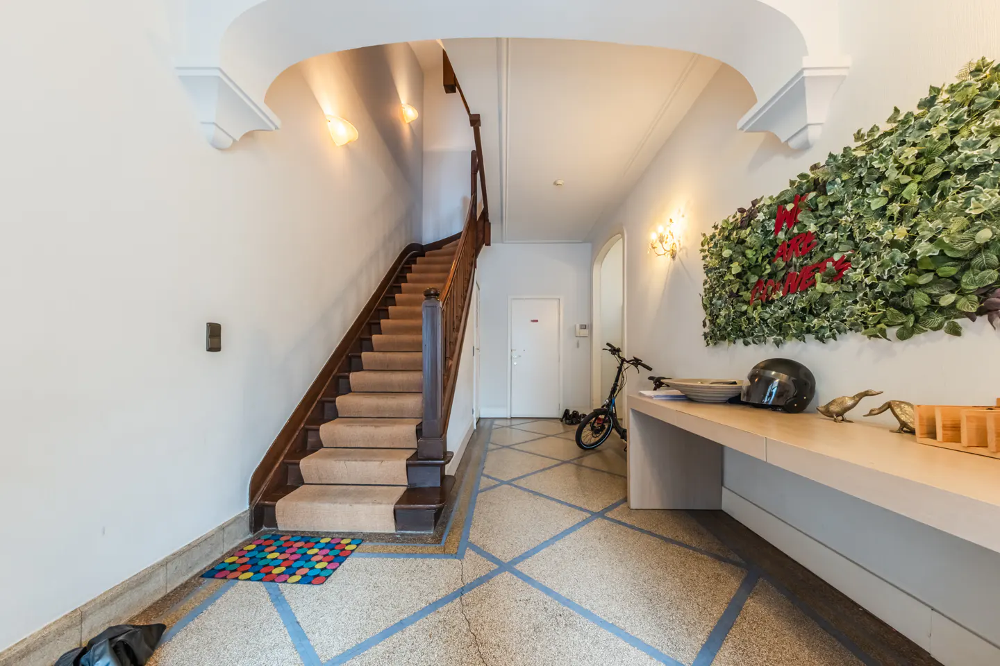 Hallway with stairs, a console table, and a bicycle. The walls are white, and the floor is speckled with blue lines. A green wall decoration is on the right.