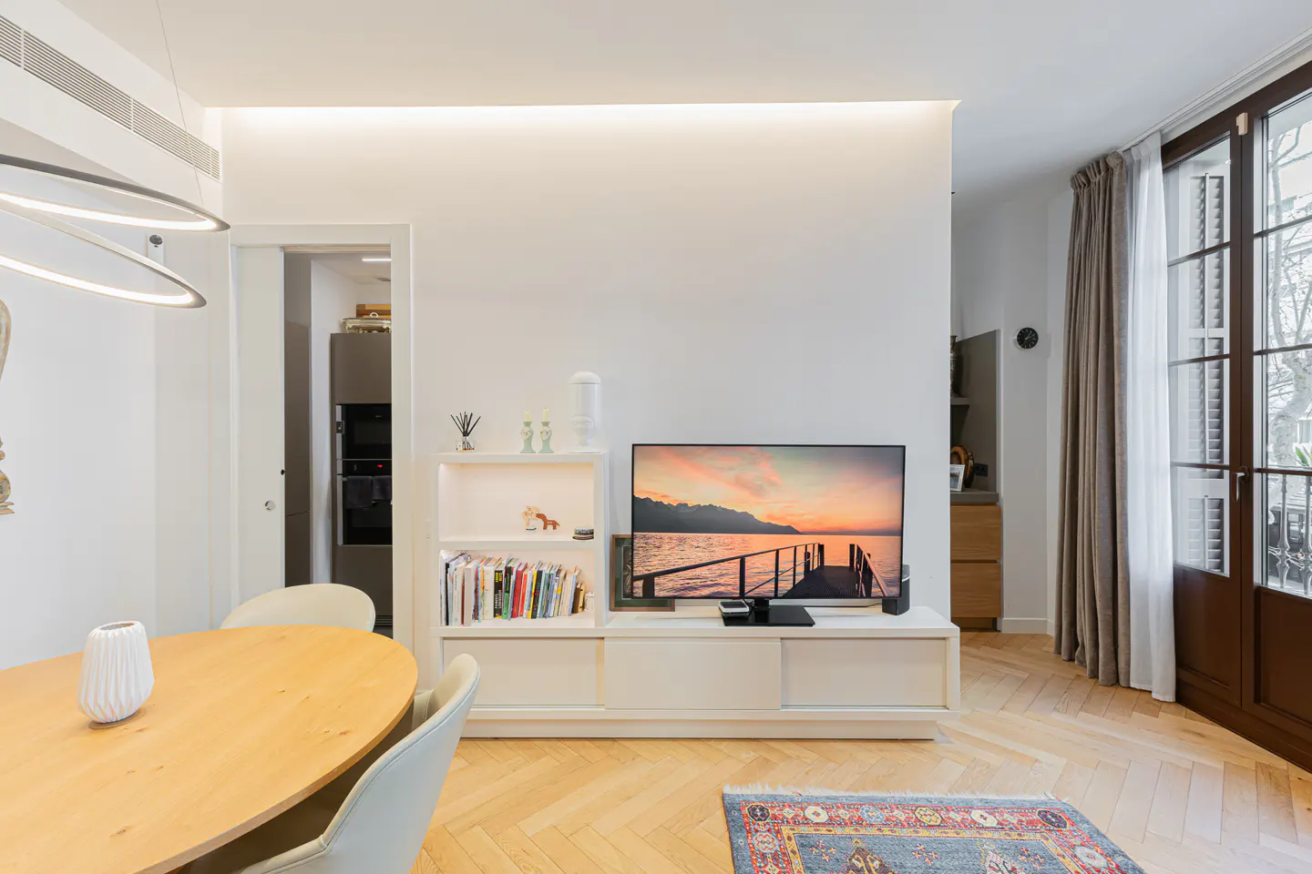 Modern living room with a light wood oval table, white media console, TV showing a sunset, and herringbone wood floors.