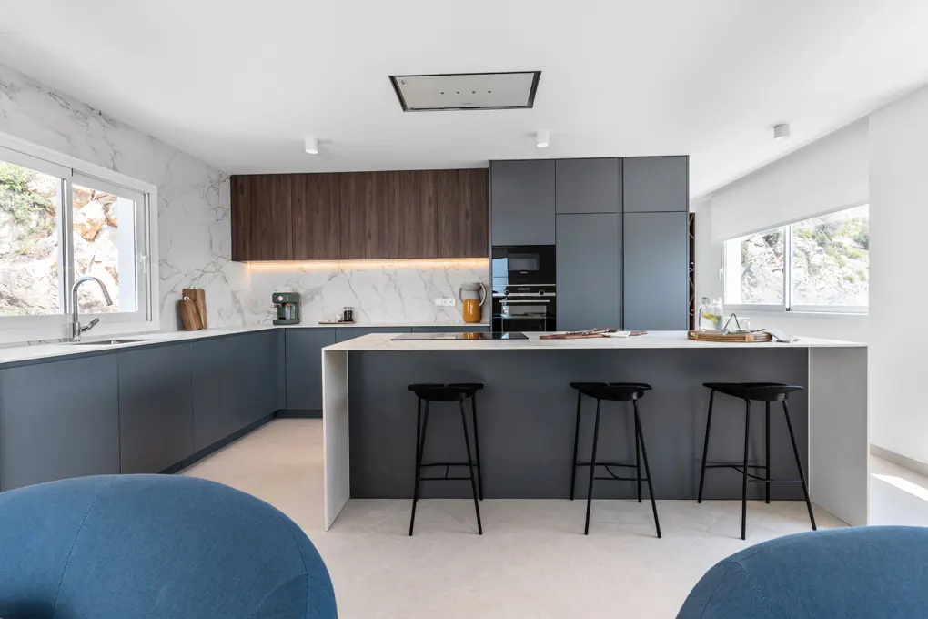 Modern kitchen with gray cabinets, white countertops, and a central island with three black stools. Marble backsplash and natural light from windows.