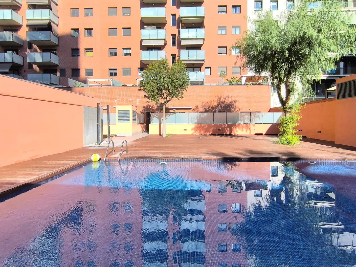 Outdoor pool with blue tile reflects a tall orange building. Wooden deck surrounds the pool with trees and a yellow ball nearby.