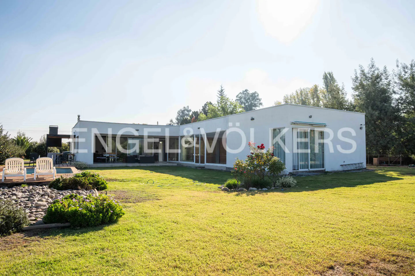 Modern white house with a green lawn, pool, and patio furniture. Engel & Völkers logo is superimposed on the house.