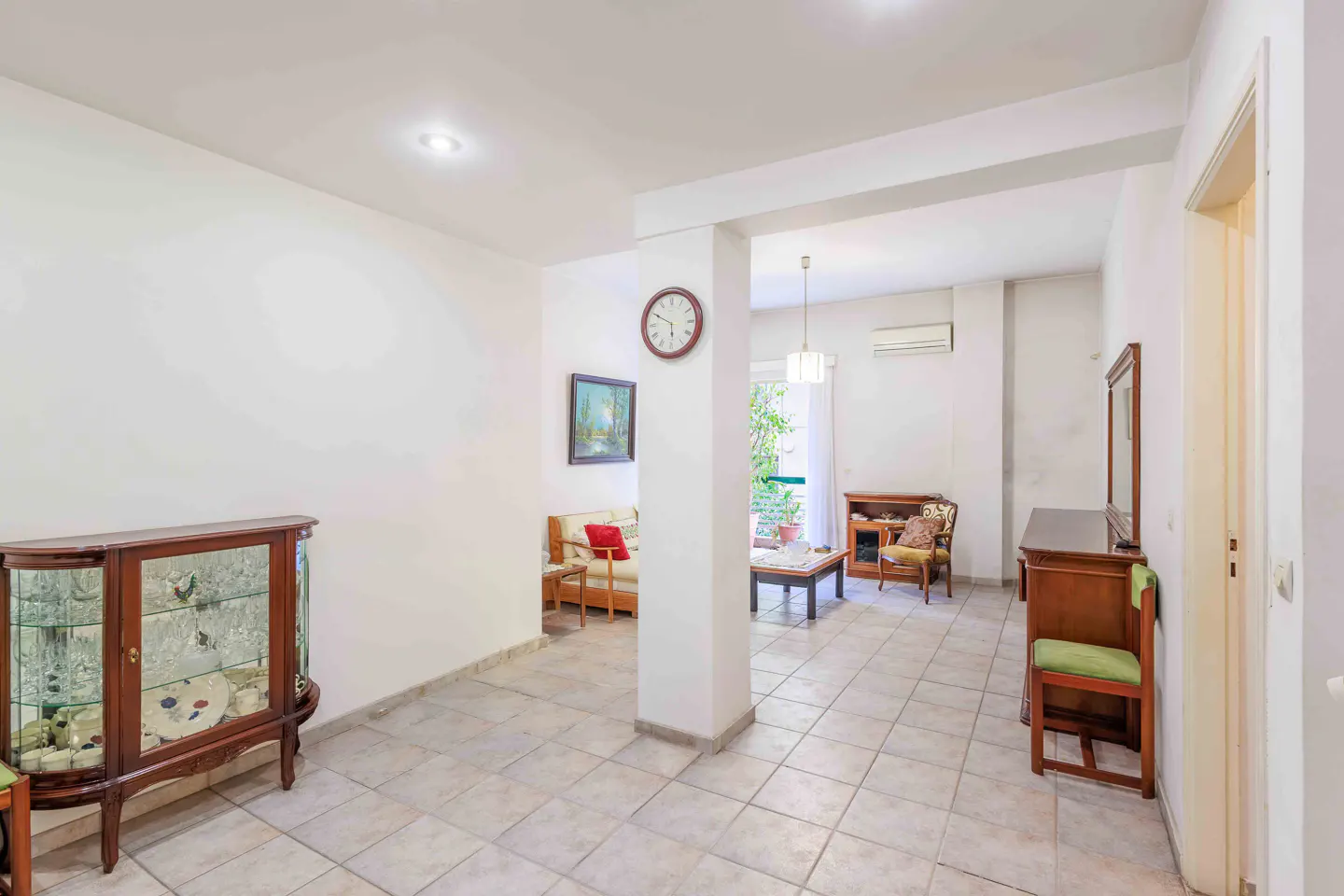 Bright, open-plan living area with tiled floor, white walls, and wooden furniture. A glass cabinet sits to the left.