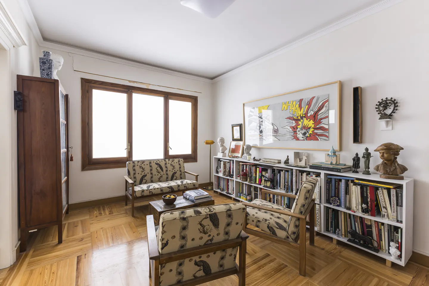 Living room with parquet floor, white walls, and a window. There is a sofa, chair, bookcase, and a wooden cabinet.