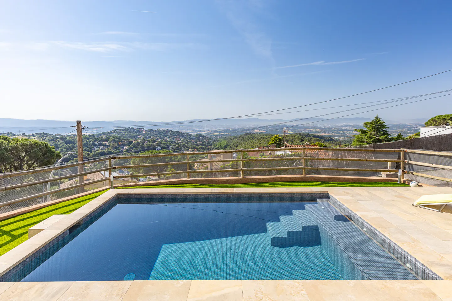 A rectangular pool with blue tiles and steps, surrounded by a wooden fence and a scenic view of green hills.