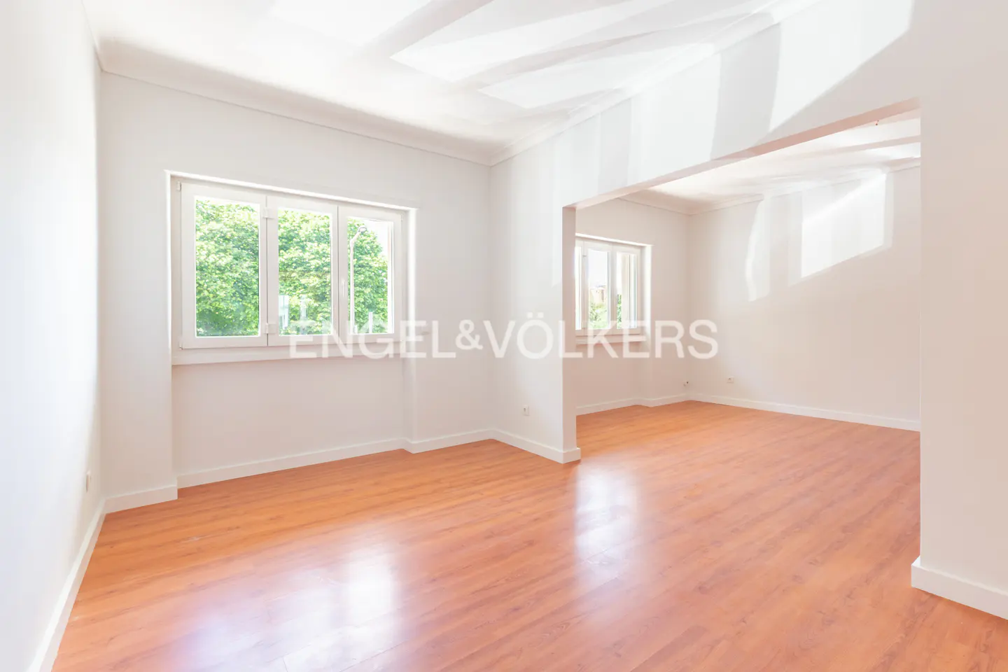 Bright, empty room with wood floors and white walls. Two windows show green trees outside. Engel & Völkers logo is visible.