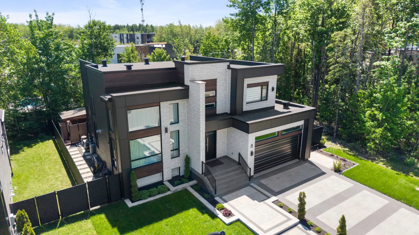Aerial view of a modern two-story home with dark wood and light brick siding, a garage, and a lush, tree-lined backyard.