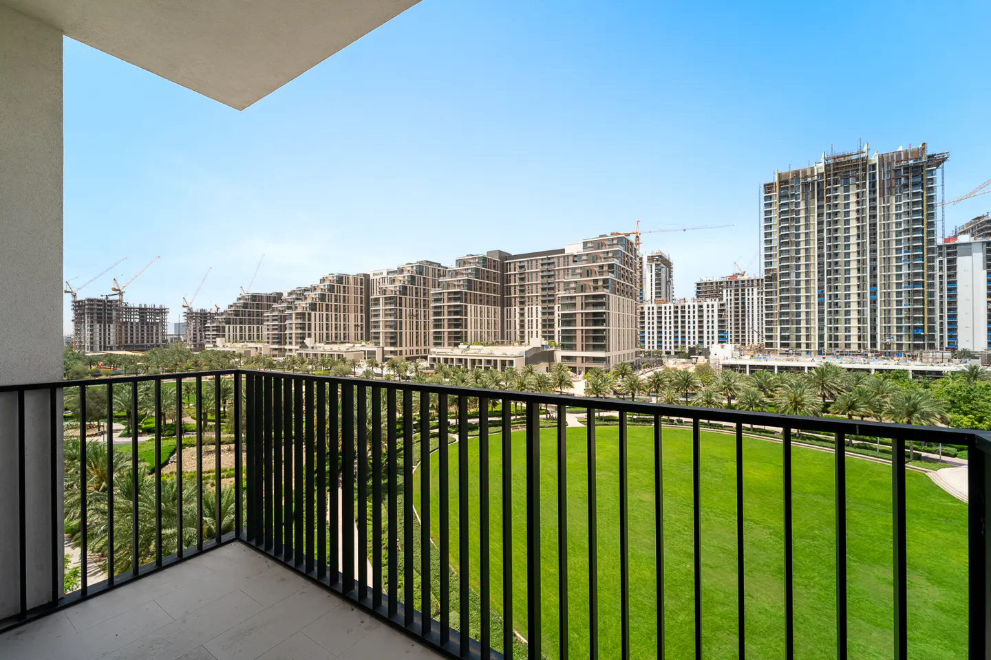 View from a balcony with black railings overlooking a green lawn, palm trees, and modern buildings under a blue sky.
