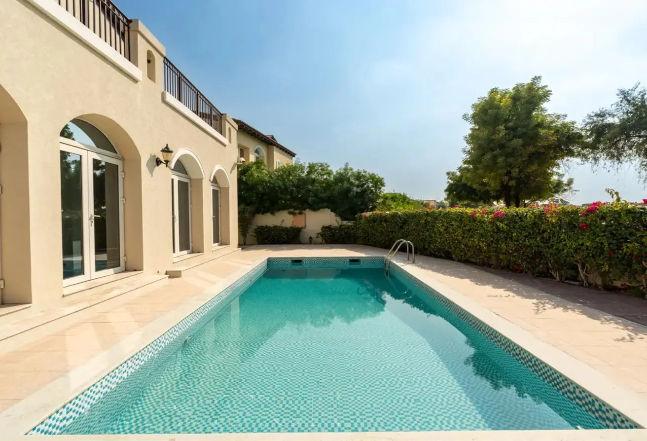 A rectangular pool with blue mosaic tiles sits beside a beige house with arched windows and a green hedge.