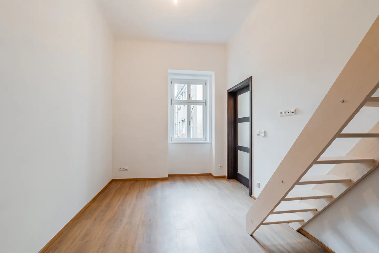 Empty room with light wood floors, white walls, a window, a dark-framed door, and a light wooden staircase on the right.