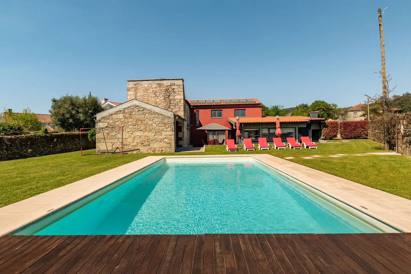 Outdoor pool with turquoise water, surrounded by green lawn, red lounge chairs, and a stone house under a clear blue sky.