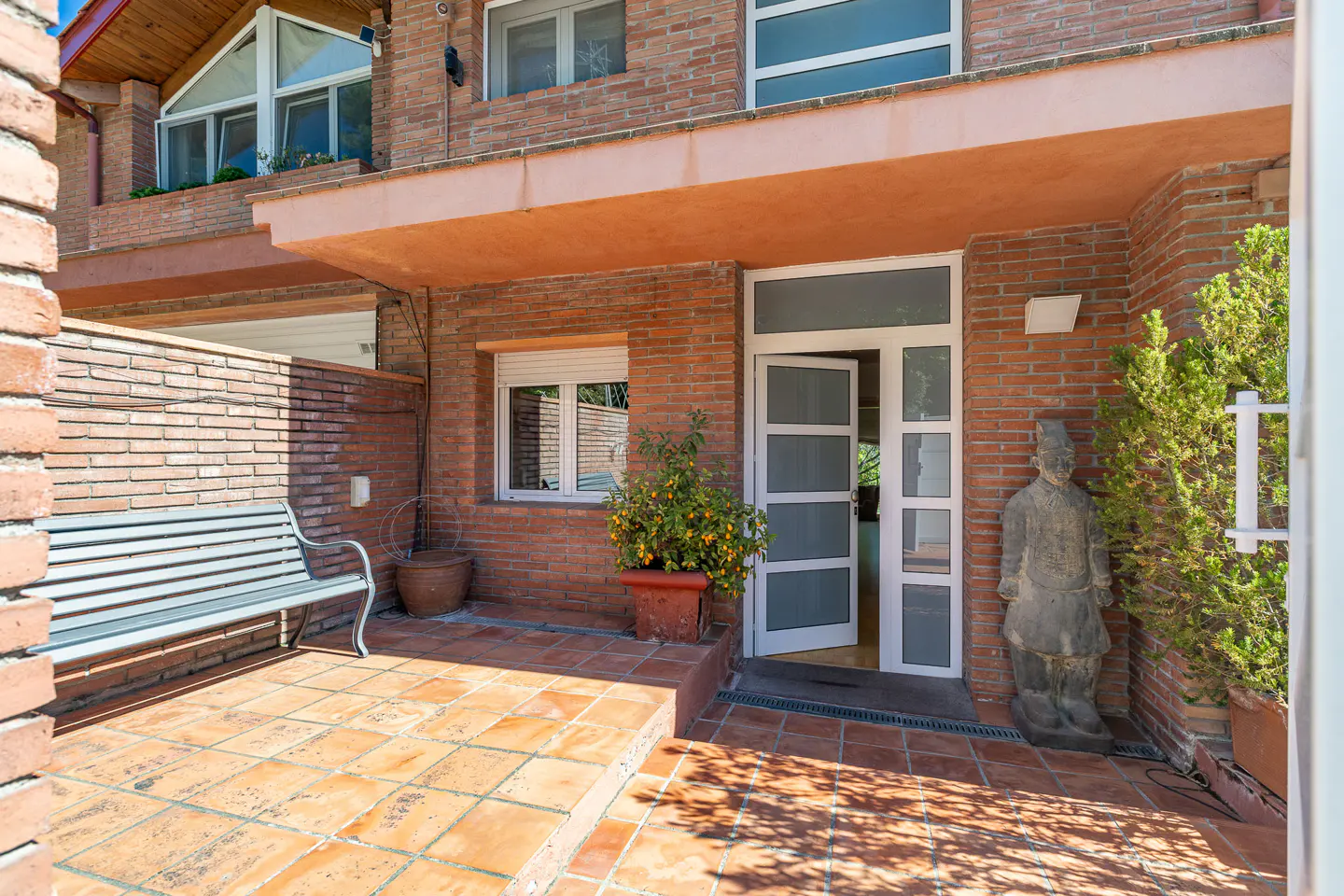 Brick house entrance with open white door, terracotta tiled patio, bench, potted plants, and a statue.
