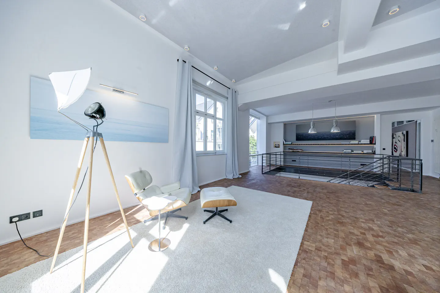 Bright, modern living room with white walls, wood floors, and a white rug. A white chair and ottoman sit near a window with light blue curtains. A tripod lamp stands nearby.