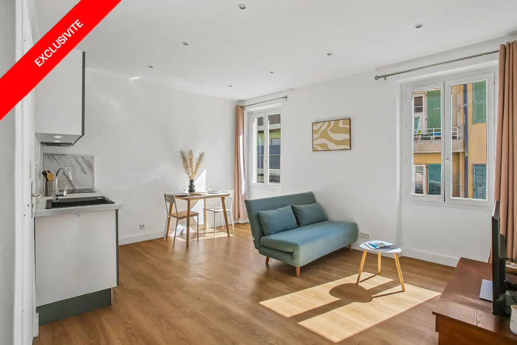 Bright studio apartment with wood floors, white walls, and natural light. A blue sofa, small table, and kitchen area are visible. "Exclusivite" banner in the corner.