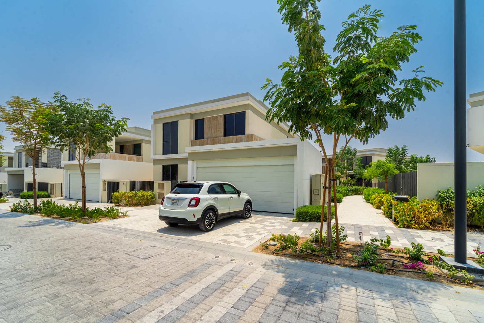 White SUV parked in the driveway of a modern two-story beige villa with a garage, surrounded by trees and paved walkways. [AI-generated text]