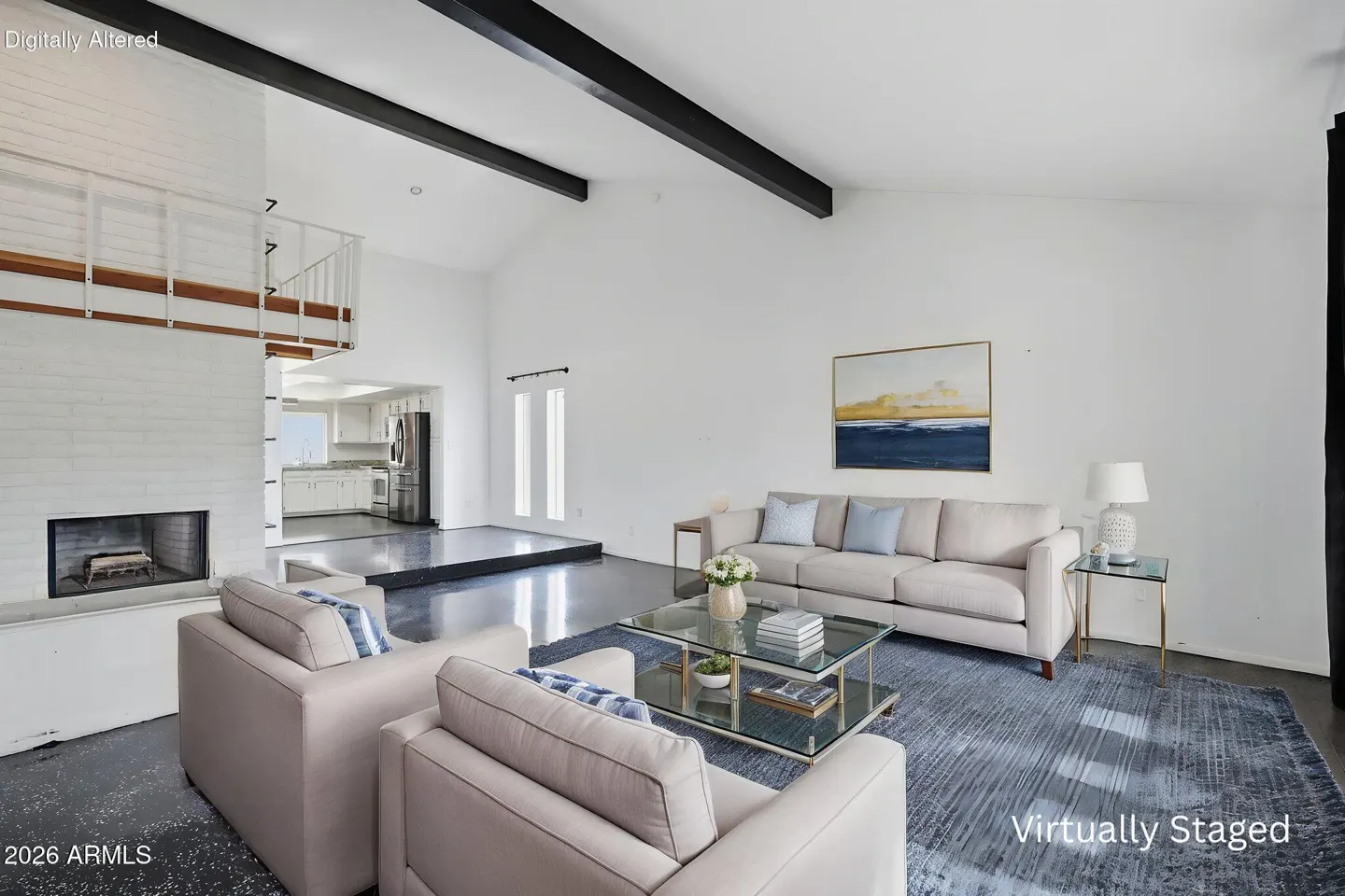 Bright, airy living room with white walls, black beams, and a fireplace. Beige sofa and chairs sit on a blue rug. Kitchen visible in the background.