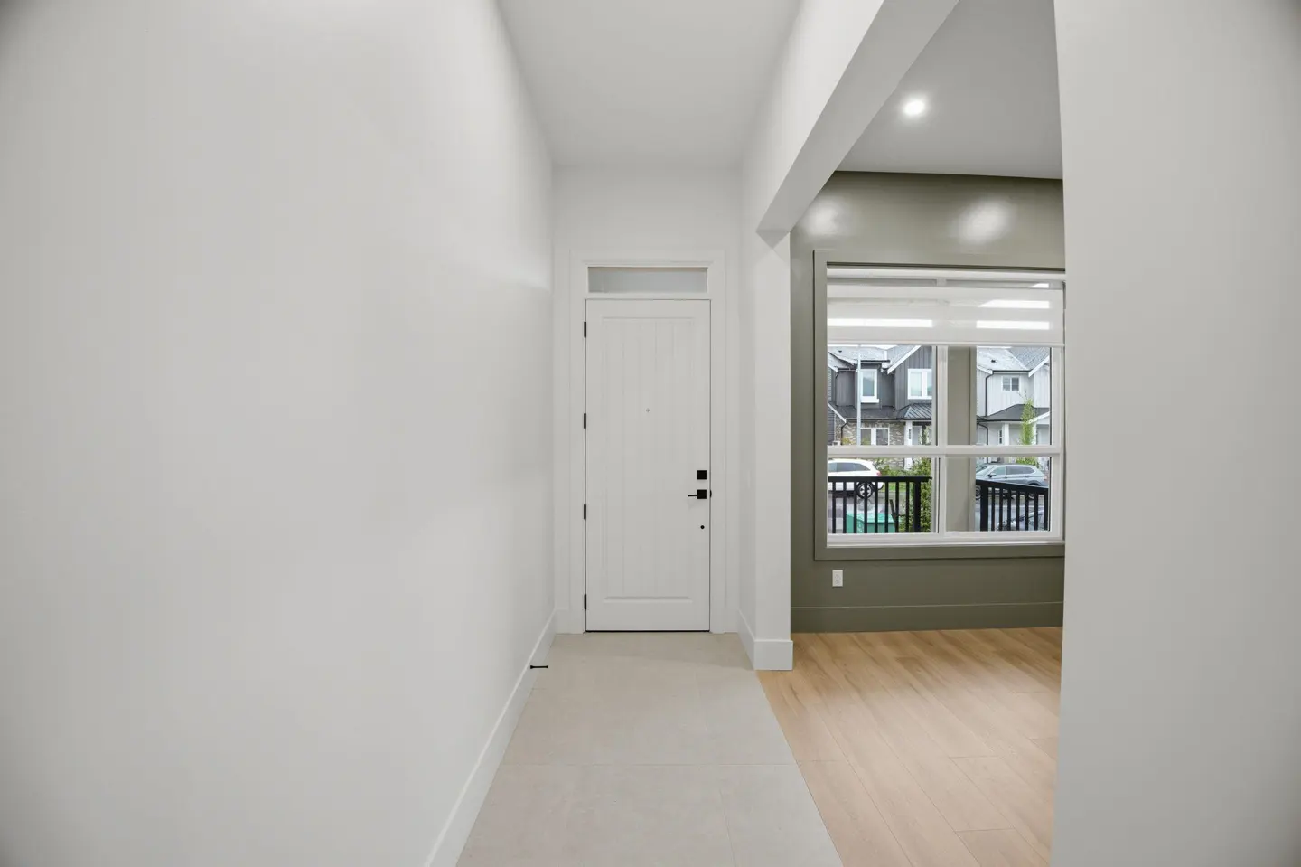 Bright entryway with white walls, a white front door, and light wood floors. A large window offers a view of houses outside.