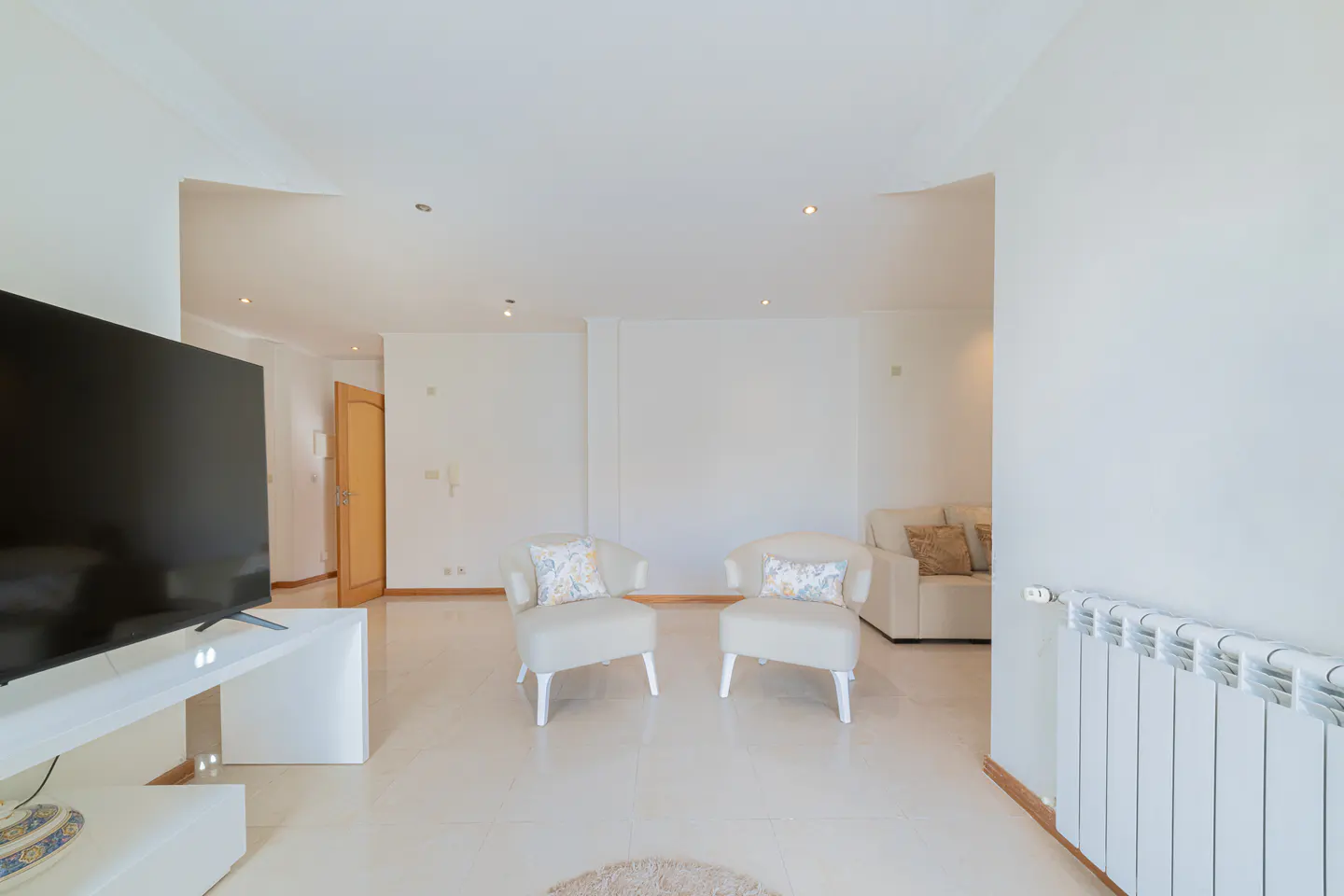 Bright living room with white walls, tile floors, and recessed lighting. Two white armchairs face a large TV. A white sofa is visible in the background.