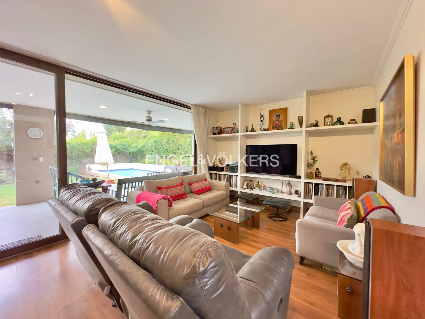 Living room with grey leather sofas, a TV on a white shelving unit, and a glass door opening to a backyard pool area.