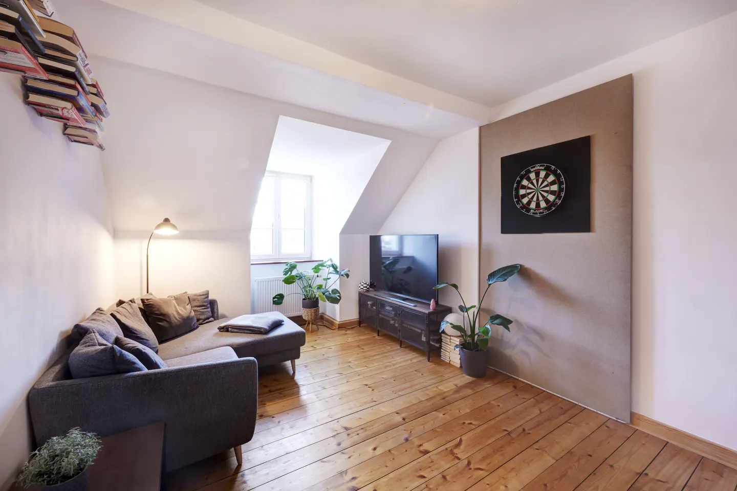 Attic living room with wood floors, gray sofa, TV, and dartboard. Books are stacked on the wall. A window provides natural light.