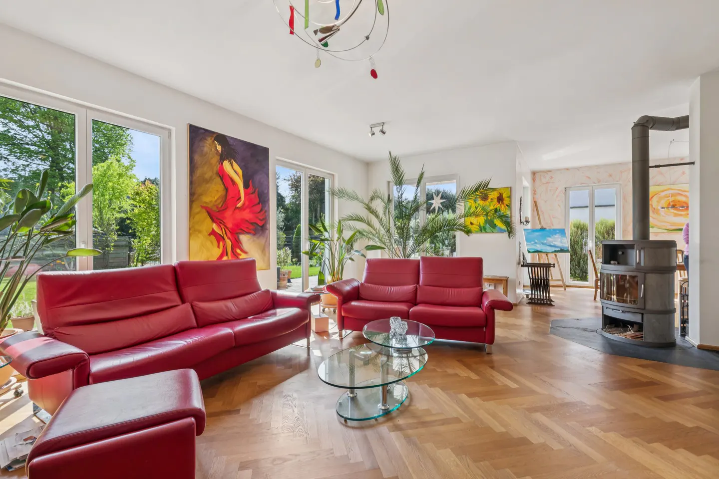 Bright living room with red leather sofas, glass tables, and a wood-burning stove on a herringbone wood floor. Large windows and paintings add color.