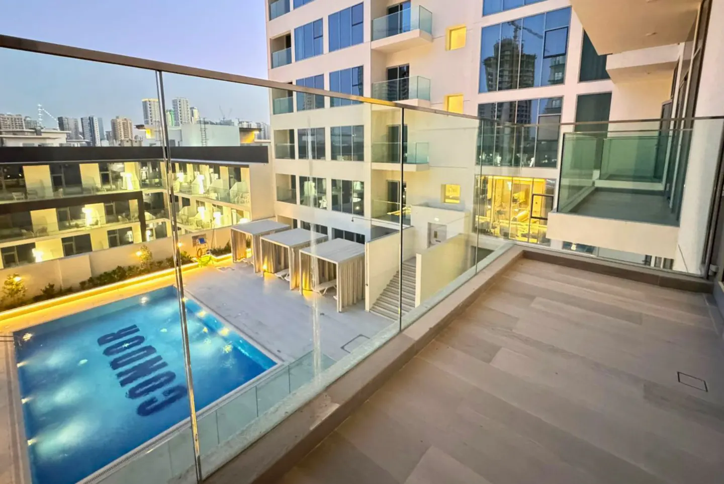 View from a balcony with glass railings overlooking a pool with "CONDOR" written in it, cabanas, and city buildings.